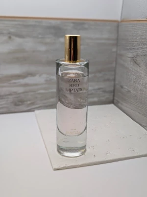 Zara Red Temptation Eau de Parfum 100ml 90% kvar - Zara Red Temptation Eau de Parfum i en elegant, cylinderformad glasflaska med guldfärgad spraykork. Flaskan är genomskinlig och parfymen har en lätt rosa ton. Stilen är minimalistisk och lyxig, perfekt för dig som gillar trendiga dofter.