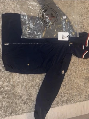 Marinblå vindjacka från Moncler - Snygg marinblå vindjacka från Moncler med huva och dragkedja framtill. Jackan har Moncler-logga på ärmen och vita samt röda ränder på huvan för en sportig look. Tillverkad i lätt och tåligt material, perfekt för blåsiga dagar.