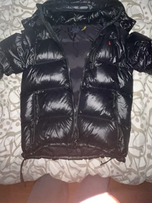 Svart pufferjacka från Polo Ralph Lauren - Svart glansig pufferjacka från Polo Ralph Lauren dragkedja framtill. Jackan har två stora fickor med dragkedja och en liten röd logga på bröstet. Perfekt för lite kyliga sommardagarna och riktigt snygg stil.