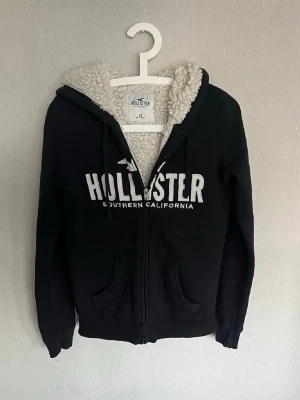 Svart Hollister hoodie med teddyfoder - Mysig svart hoodie från Hollister med dragkedja och vit logga på bröstet. Insidan av huvan är fodrad med vitt teddyfoder för extra värme. Två fickor framtill och justerbar huva med snören. Perfekt för kalla dagar och chill stil. Skicket är bra, några små trådar som är synliga i huvan men inget man tänker på. Säljer den då den inte riktigt passade som jag hade tänkt. Pris är diskuterbart så var inte rädd att fråga!🙌