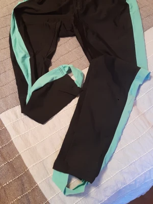 Retro aerobics-style från 1980s... en svarta träningsbyxor med turkosa detaljer - Return to the 1980s aerobics style... Säljer ett par svarta träningsbyxor med turkosa ränder längs sidorna och fotstroppar. Byxorna har en smal passform och är tillverkade i ett stretchigt syntetmaterial som passar perfekt för sport och träning.