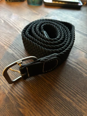 Svart flätat bälte 110cm - Snyggt svart flätat bälte med silverfärgat spänne och detaljer i skinnimitation. Perfekt för att lyfta vilken outfit som helst och passar till både jeans och byxor. Enkel att justera tack vare det flexibla materialet.