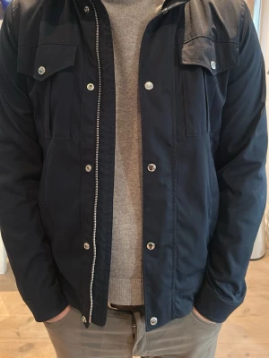 Mörkblå jacka från J.Lindeberg - Stilren mörkblå jacka från J.Lindeberg med silverfärgade knappar och dragkedja. Jackan har två bröstfickor med lock och knapp samt klassisk krage. Perfekt för dig som gillar en clean och modern look.