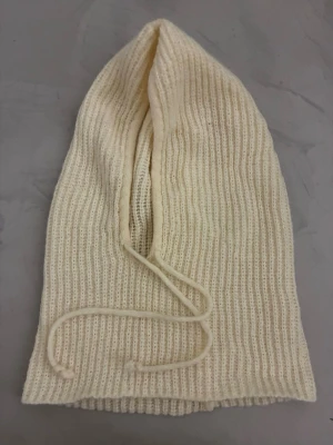Ribbstickad balaklava i offwhite - Säljer en ribbstickad balaklava i offwhite med dragsko framtill. Mössan har en lång, avlång form och är tillverkad i ett mjukt, stickat material. 