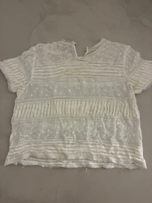 Vit spetsblus med broderi från Viscose - Söt vit blus i tunn spets med broderade detaljer och små prickar. Blusen har korta ärmar och en  rund hals med liten v-ringning.