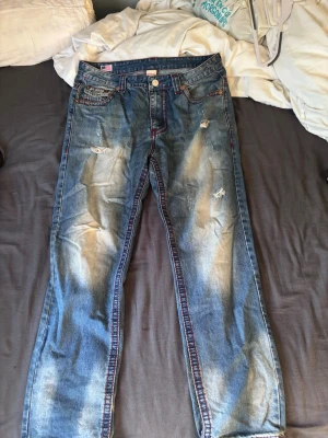 True Religion slitna blå jeans - Säljer ett par blå jeans från True Religion med slitningar och distressed detaljer på benen. En av knapparna på bakfickan har ramlat av.