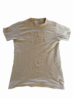 Beige Lacoste t-shirt  - Snygg beige t-shirt från Lacoste med upphöjd krokodil och text på bröstet. Klassisk rund halsringning och korta ärmar. Tillverkad i mjuk bomull och har en clean, stilren look som passar perfekt till jeans eller shorts.