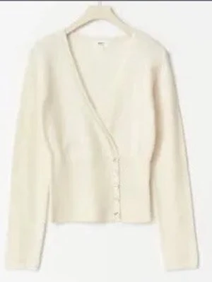 Krämvit kofta med v-ringning - vit kofta med djup v-ringning och knappar framtill. Koftan har lång ärm och är ribbstickad nedtill för en snygg passform. Materialet ser mjukt och fluffigt ut, perfekt för lager på lager. Enkel och stilren design som passar till det mesta.