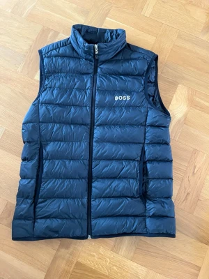 Svart dunväst från BOSS - Snygg svart dunväst från BOSS med quiltad design och hög krage. Västen har dragkedja framtill, två sidofickor och diskret BOSS-logga på bröstet. Perfekt för lager-på-lager och ger en clean look till din outfit.