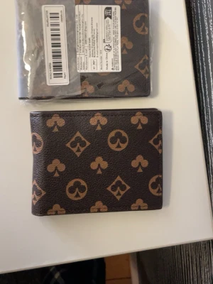 Brun plånbok med spelkortsmönster - Snygg brun plånbok i konstläder med spelkortsinspirerat mönster i beige. Plånboken har flera fack för kort, ett fönster för ID och en liten myntficka med dragkedja. Perfekt för dig som vill ha en unik och stilren accessoar. Du köper du oöppnade😊