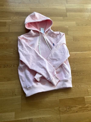 Ljusrosa hoodie från Polo Ralph Lauren - Snygg ljusrosa hoodie från Polo Ralph Lauren med klassisk huva och dragsnören. Broderad blå logga på bröstet och känguruficka framtill. Mjuk bomullskänsla och ribbade muddar vid ärmslut och nederkant. Perfekt för en chill och stilren look.