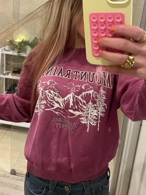 Lila sweatshirt Mount Rainier print - Mörk Lila sweatshirt med vitt Mount Rainier-tryck framtill. Motiv med berg, träd och texten 'National Forest'. Mjuk bomullskänsla och avslappnad passform med rund hals och ribbade muddar. Köpt på Hm för ett tag sen men har bara använt några få gånger. Skriv gärna vid frågor💕
