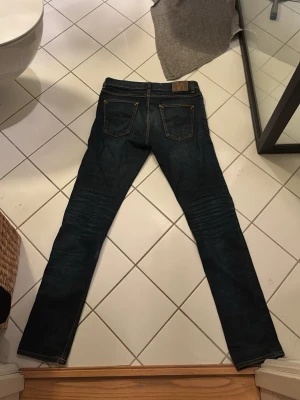 Mörkblå jeans från Nudie Jeans - Säljer ett par mörkblå jeans från Nudie Jeans med klassisk femficksmodell och raka ben. Jeansen har kontrastsömmar i orange och Nudie-logga på bakfickan. Tillverkade i slitstarkt denimtyg, perfekta för en avslappnad och stilren look.