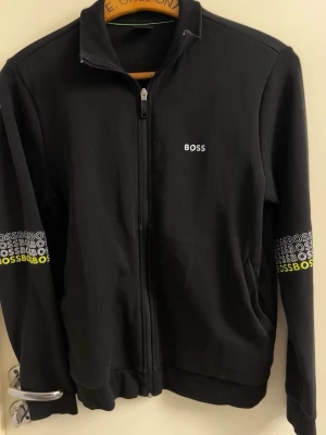 BOSS Tröja - Snygg svart ziptröja från BOSS med vit logga på bröstet och upprepade BOSS-tryck i vitt och gult på ärmarna. Tröjan har hög krage, hel dragkedja och ribbade muddar. Perfekt för en sportig och avslappnad stil. KVITTO OCH BEVIS PÅ KÖP FINNS VID FÖRFRÅGAN, pris kan diskuteras vid snabb affär. STORLEK S/M