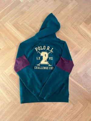 Grön Ralph Lauren hoodie Challange cup - Säljer denna snygga RL hoodien i grönt st M!🔥😎 Hoodien har stilrena detaljer med en snygg logga på framsida vänster bröst och en stilren design på ryggen! Ärmarna har vinröda delar för att sticka ut och ge det lilla extra den behöver! Skick - 9/10, knappt använd, märks på det exemplariska skicket! Storlek - M, kan lika gärna passa L! Mått - Bröstbredd 53, längd 69, armlängd 67. Tveka inte att höra av er vid frågor eller funderingar!