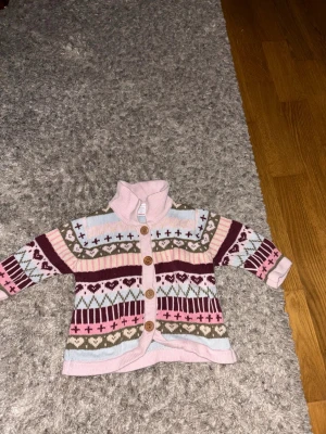 Mönstrad stickad kofta i pastellfärger - Säljer en söt stickad kofta med knappar framtill och hög krage. Koftan har ett färgglatt mönster i rosa, vinrött, ljusblått, beige och vitt med hjärtan och kors. Perfekt för dig som gillar mysiga och färgglada plagg.