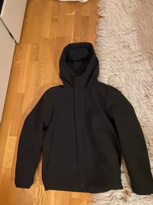 Svart jacka från Zara - Säljer en stilren svart vindjacka med huva och dragkedja framtill. Jackan har en minimalistisk design, raka ärmar och justerbar nederkant. Materialet ser ut att vara vattenavvisande syntet, perfekt för blåsiga dagar. Inga synliga loggor eller märken.