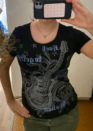 Svart t-shirt med gitarrtryck - Cool svart t-shirt med stort tryck av en elgitarr och stjärnor på framsidan. Texten 'Rock' och 'Rebel' syns i blått och vitt. T-shirten har rund hals och korta ärmar, perfekt för dig som gillar rockig stil.