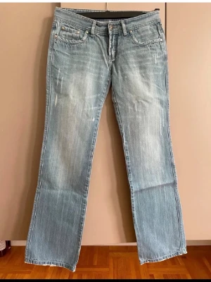 Ljusblå  jeans med slitningar - Säljer ett par ljusblå jeans med slitningar framtill och coola gula sömmar på bakfickorna. Klassisk femficksmodell med knappgylf och avslappnad passform. Perfekta till sneakers eller boots för en chill look.