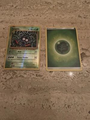 Två Pokémonkort: Tangela & Energi - Säljer två Pokémonkort: ett holografiskt Tangela-kort med grönt tema och ett grönt Grass Energy-kort. Perfekt för samlare eller dig som vill bygga din lek. Båda korten har klassisk Pokémon-baksida och färgstarka motiv.