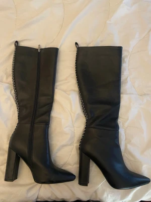 Svarta höga boots med blockklack - Snygga svarta stövlar med hög blockklack och spetsig tå. Dragkedja på insidan och dekorativ vit söm längs baksidan som ger en cool kontrast. Tillverkade i slätt konstläder. Ca 10 cm höga.