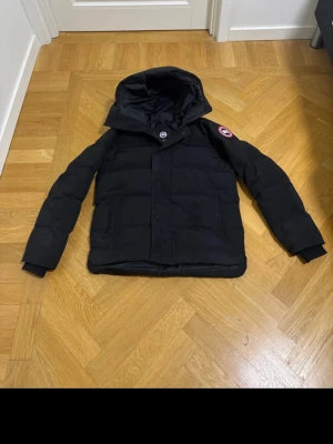Svart Canada goose Macmillan  - Snygg svart dunjacka från Canada Goose med huva och klassisk logga på ärmen. Jackan har quiltad design, dragkedja och tryckknappar framtill. Perfekt för kalla vinterdagar och riktigt varm. Passar dig som vill ha både stil och funktion.