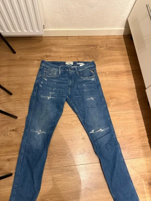 Blå slitna jeans från Replay - Säljer ett par blå jeans från Replay med slitningar och distressed detaljer på framsidan. Klassisk femficksmodell med raka ben och normal midja. Jeansen har snygga sömmar och läderpatch med logga bak i midjan. Perfekta för en avslappnad och trendig stil. Ny pris 1800kr