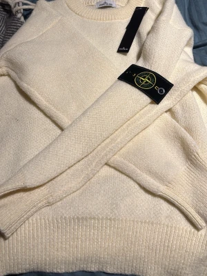 Vit stickad tröja från Stone Island - Säljer en stilren vit stickad tröja från Stone Island med klassisk rund halsringning och ribbade muddar. Tröjan har den ikoniska Stone Island-loggan på ärmen och är gjord i mjukt, tjockt material som håller dig varm. Perfekt för dig som gillar streetwear och snygga detaljer.