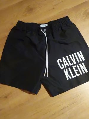 Svarta badbyxor från Calvin Klein oanvända  - Snygga svarta badbyxor från Calvin Klein med vit logga tryckt på vänster ben. De har elastisk midja och vita dragsnören för justerbar passform. Perfekta för stranden eller poolen och tillverkade i snabbtorkande material.