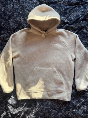 Peak Performance Hoodie - Mysig beige hoodie i fleece med stor magficka. Hoodien har en enkel och clean design utan skrikigt märke, perfekt för dig som gillar en avslappnad stil. Långa ärmar och mjukt material som håller dig varm under kyliga dagar.