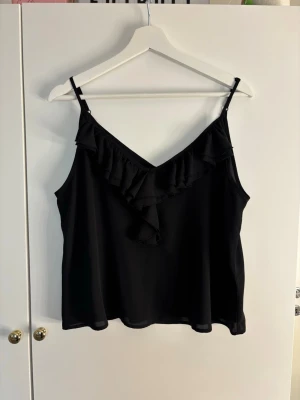 Svart linnetopp med volang från Bik Bok - Snygg svart linnetopp från Bik Bok med smala axelband och volangdetalj framtill. Toppen har v-ringning och är tillverkad i ett mjukt, lätt material som känns skönt mot huden. Perfekt att styla med jeans eller kjol för en trendig look.