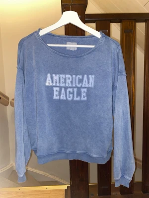 Blå sweatshirt från American Eagle - Blå sweatshirt med avslappnad passform från American Eagle. Tröjan har rund halsringning, ribbade muddar och tryckt logga i vitt framtill. Perfekt för en chill och casual stil.