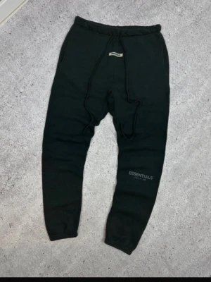 Svarta Essentials mjukisbyxor - Svarta mjukisbyxor från Essentials med tryckt logga på benet. Byxorna har resår i midjan, dragsko och muddar vid bensluten. Perfekta för en chill och avslappnad stil. Tillverkade i mjukt material för extra komfort.