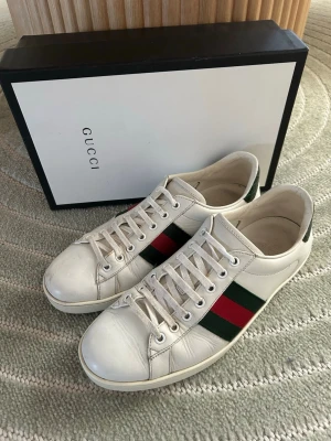 Gucci vita sneakers med röd-gröna ränder - Snygga vita sneakers från Gucci med klassiska röd-gröna ränder på sidorna. Skorna har låga skaft, vita snören och är tillverkade i läder. Perfekta för dig som vill ha en stilren och lyxig look med ikoniska detaljer.