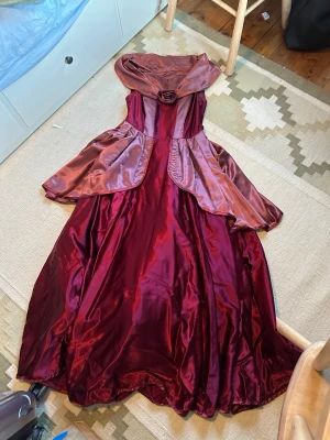 Vinröd balklänning  - Säljer mammas gamla balklänning från 1993 som endast är använd en gång. Finns bara ett exemplar då hon designade den själv! Kan gå ner i pris vid snabb affär💕💕Vinröd långklänning i glansigt material med bred volangkrage och dekorativ ros framtill. Klänningen har utställd kjol och volangdetaljer över axlarna, vilket ger en elegant look. Perfekt för bal eller festliga tillfällen.