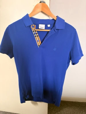 Blå pikétröja från Burberry  - Snygg blå pikétröja från Burberry med klassiskt rutigt mönster vid knäppningen och blå broderad logga på bröstet. Kortärmad modell i bomull med två blå knappar och krage. Perfekt för en stilren och avslappnad look. Stealerbjudande på 699 kr gäller fram till onsdag 23:59