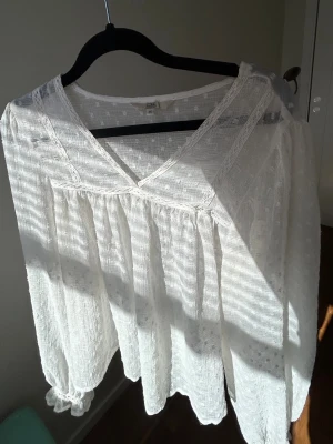 Vit bohemisk blus med spetsdetaljer - Luftig vit blus i bohemisk stil med v-ringning och spetsdetaljer längs halsringningen. Blusen har ett lätt transparent tyg med diskret struktur och långa, lösa ärmar med resår vid handlederna. Perfekt för en avslappnad och trendig look🥂💗