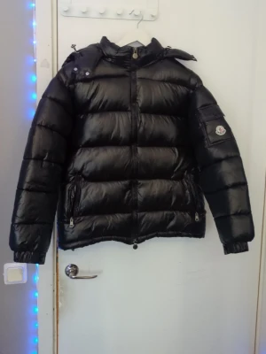 Svart dunjacka från Moncler - Snygg svart dunjacka från Moncler med glansig finish, huva och två dragkedjeförsedda fickor. Jackan har en patch med Moncler-loggan på ärmen och grov dragkedja framtill. Fungerande nfc till Moncler hemsidan.