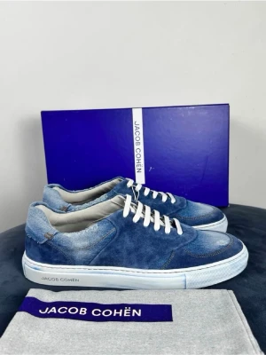 Blå sneakers från Jacob Cohën - Snygga blå sneakers från Jacob Cohën i en mix av mocka och denim. Skorna har vita snören, vit sula med logga och coola kontrastsömmar. Perfekta för dig som gillar en avslappnad men trendig stil. Kommer med originalkartong och dustbag.