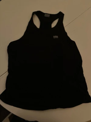 Svart träningstopp från MM Sports - Snygg svart träningstopp från MM Sports med brottarrygg och logga både framtill och nertill. Perfekt för gymmet eller andra sportaktiviteter. Tillverkad i mjukt och stretchigt material som ger bra rörelsefrihet.