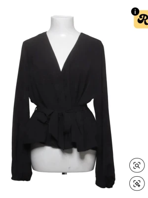 Svart omlottblus  - Stilren svart omlottblus med djup V-ringning och knytskärp i midjan som ger en snygg siluett. Blusen har långa ärmar och är tillverkad i ett mjukt, lätt tyg som faller fint. Använd någon enstaka gång och i bra skick!! 