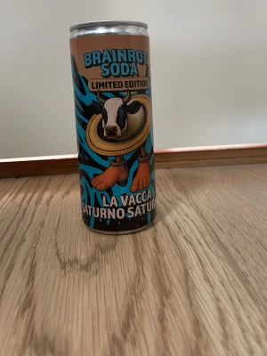 Brainrot Soda limited edition burk - Cool limited edition burk från Brainrot Soda med färgglad design och motiv av en ko med ring runt halsen. Burken har blå och bruna detaljer samt texten 'La Vacca Saturno Saturn'. Perfekt som samlarpryl eller dekoration. 1000 i hela världen