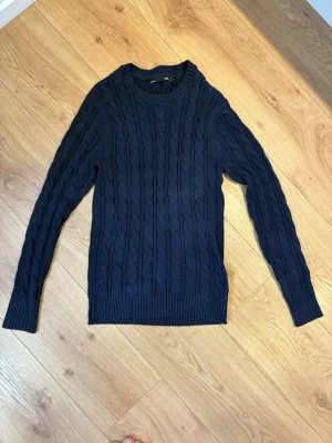 Mörkblå kabelstickad tröja  - Säljer en mörkblå kabelstickad tröja från 157 med rund halsringning och ribbade muddar vid ärmslut och nederkant. Tröjan har ett klassiskt flätmönster och är perfekt för dig som gillar en stilren och enkel look.