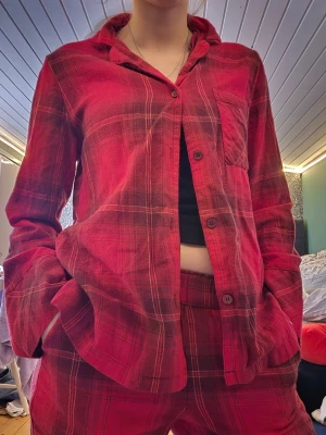 Röd- och svartmönstrad pyjamas - Mysig pyjamas i rött och svart rutmönster med skjortkrage och knappar framtill. Setet består av långärmad överdel med bröstficka och matchande byxor med resår i midjan. Perfekt för chillkvällar hemma.