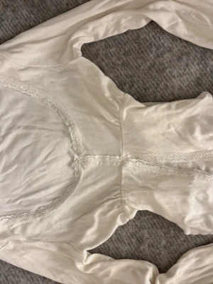 Vit långärmad topp med spetsdetaljer - Söt vit långärmad topp med spets längs urringningen och framtill. Modellen har en lätt rynkad midja vilket ger en fin silhuett och volangig nederkant. Perfekt för en romantisk och feminin stil. Tunt och mjukt material. OBS Sitter ej ihop över bål (perfekt om man har navelpiercing). Jättefint skick och endast använd fåtal gånger mycket försiktigt