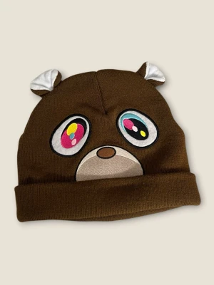 Brun kanye west mössa med öron - Säljer en brun stickad mössa med uppvikt kant och två små öron på toppen. Mössan är gjord i mjukt material och har en enkel, lekfull design som sticker ut. Perfekt för dig som vill ha något gulligt och annorlunda till hösten eller vintern.