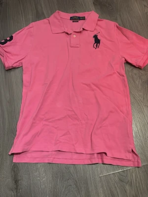 Rosa pikétröja från Polo Ralph Lauren - Säljer en klassisk rosa pikétröja från Polo Ralph Lauren i storlek XS/S. Tröjan har krage, knappar vid halsen och den ikoniska loggan broderad på bröstet. Tillverkad i mjuk bomull och har korta ärmar med en siffra på höger ärm.