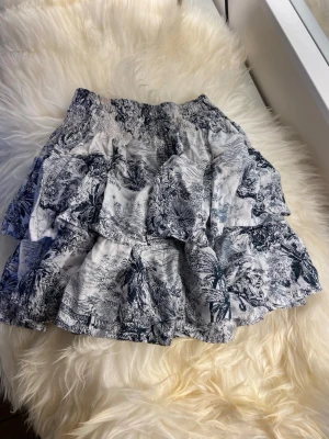 Vit volangkjol med blått mönster - Kort volangkjol i vitt med blått blommigt och naturinspirerat mönster. Kjolen har flera lager volanger och resår i midjan för en bekväm passform. Perfekt för en somrig och avslappnad stil.