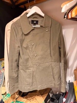 Olivgrön dubbelknäppt rock från H&M - Snygg olivgrön rock från H&M med dubbelknäppning och bred krage. Rocken har två framfickor, dekorativa sömmar och en knappdetalj bak i midjan. Tillverkad i ett mjukt bomullsliknande material och har en avslappnad passform.