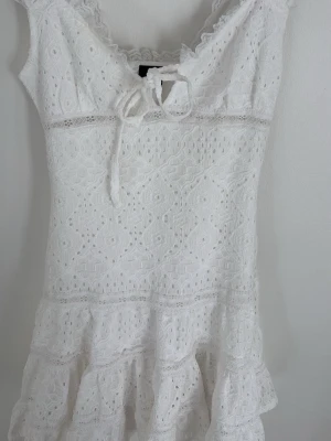 Vit spetsklänning med volang - Söt vit klänning i spets med volangkant nedtill och knytning vid urringningen. Ärmlös modell med v-ringad hals och fina spetsdetaljer. Perfekt för sommarens alla tillfällen.